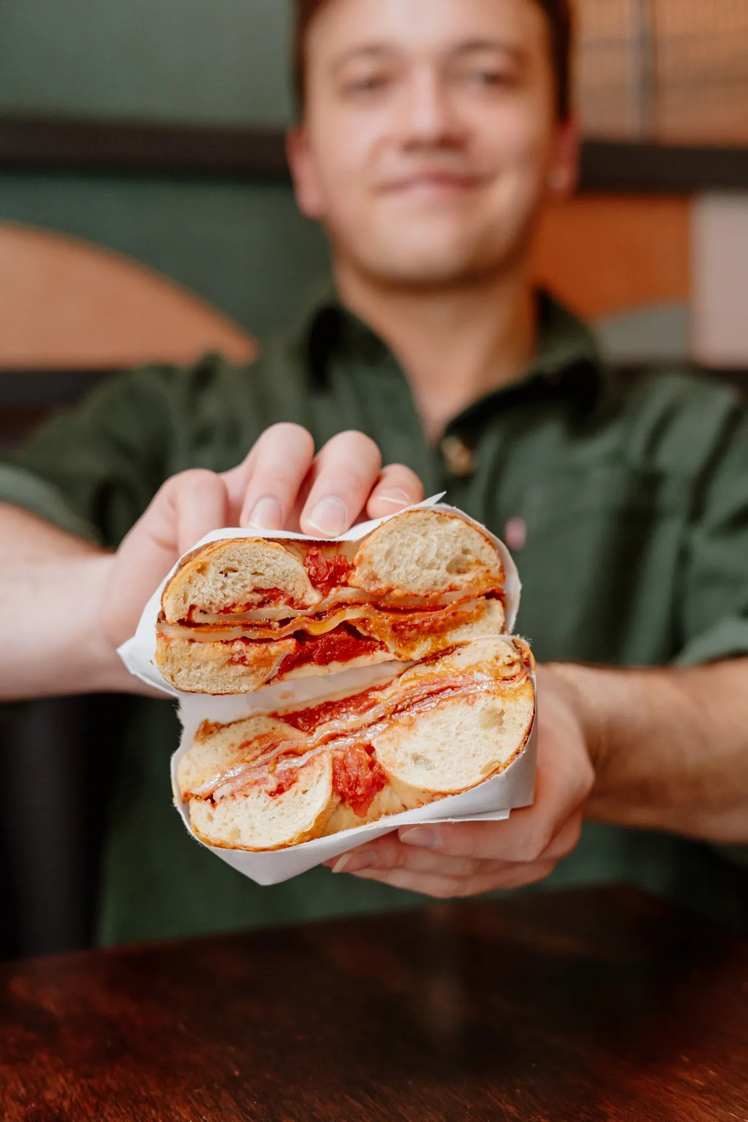 Smiling man in a green shirt holding up a halved bagel sandwich layered with tomato sauce and melted cheese