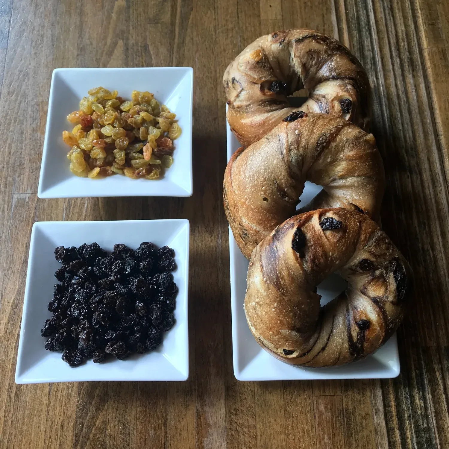 Three cinnamon raisin bagels beside square white dishes of golden and dark raisins on a wooden table