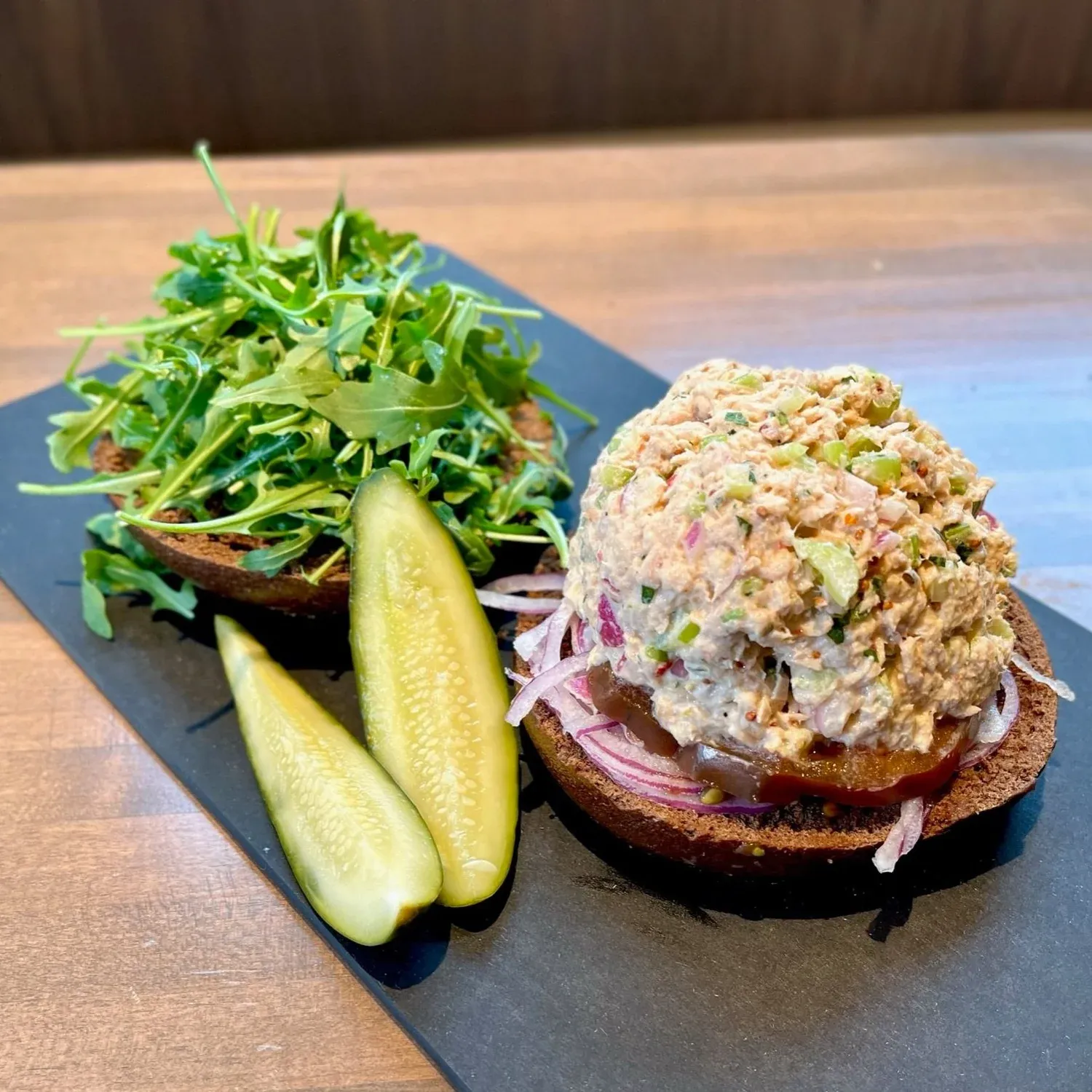 Open-faced pumpernickel bagel topped with tuna salad, tomato and red onion, with arugula and pickle spears on a slate board