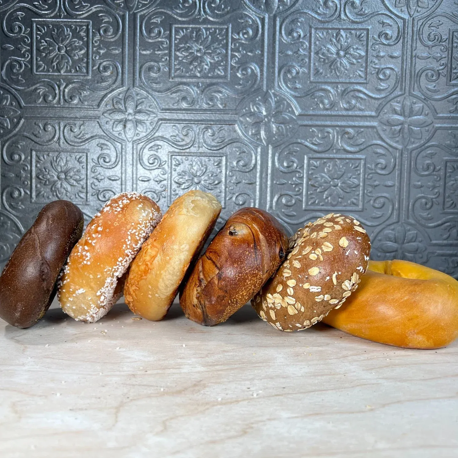 Six bagels leaning in a row against an embossed gray tin wall: pumpernickel, salt, plain, cinnamon raisin, oat and egg