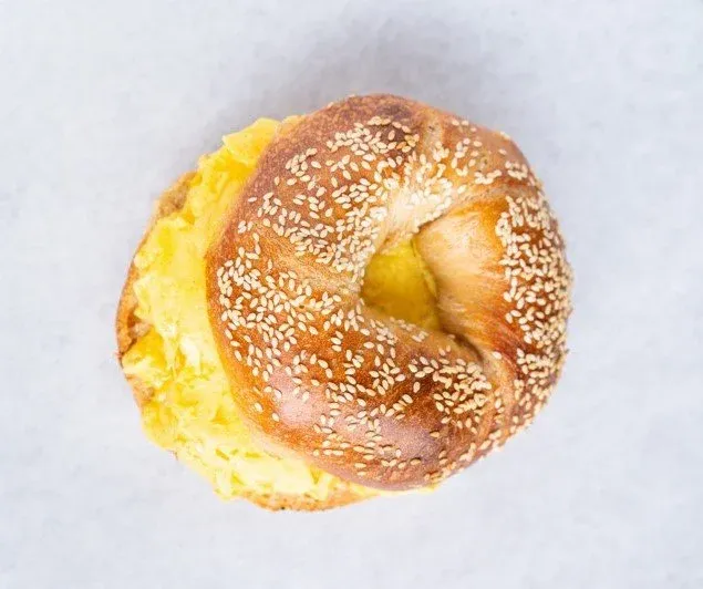 Overhead view of a sesame bagel sandwich with scrambled egg spilling out one side on white paper