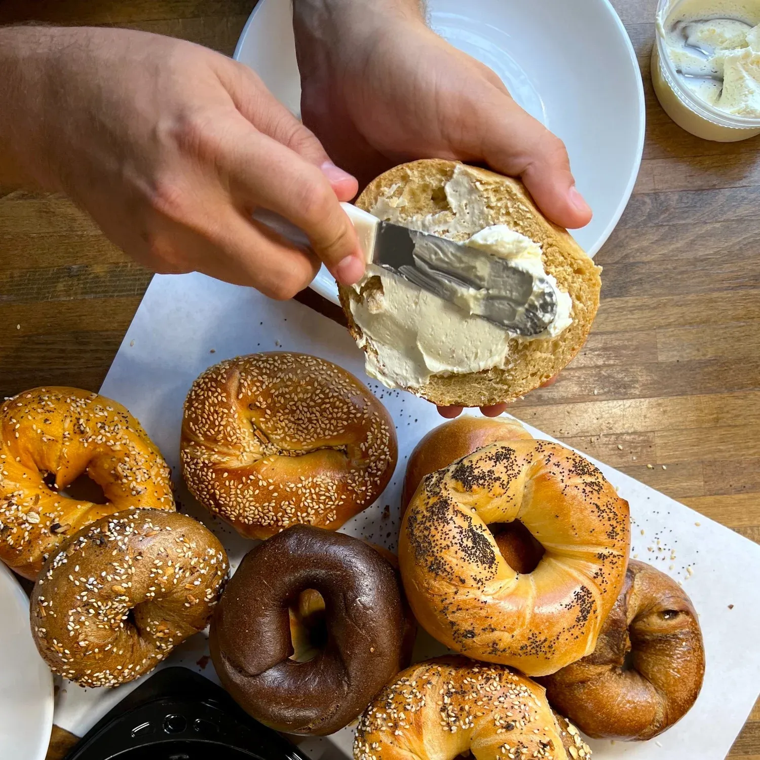 Hands spreading cream cheese on a bagel half above a tray of sesame, everything, poppy, pumpernickel and raisin bagels