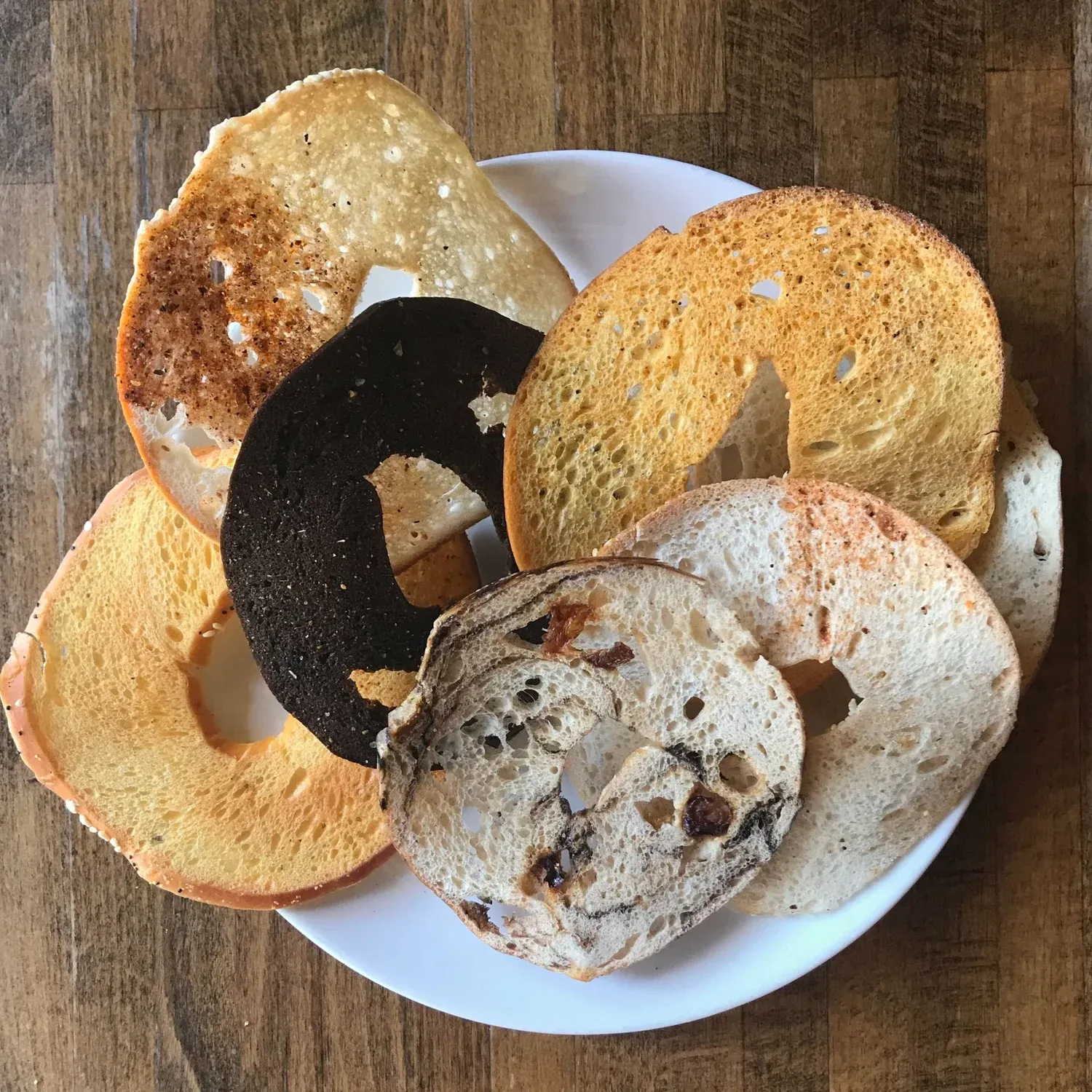 Thin crisp bagel chips in several varieties, including pumpernickel and cinnamon raisin, stacked on a white plate