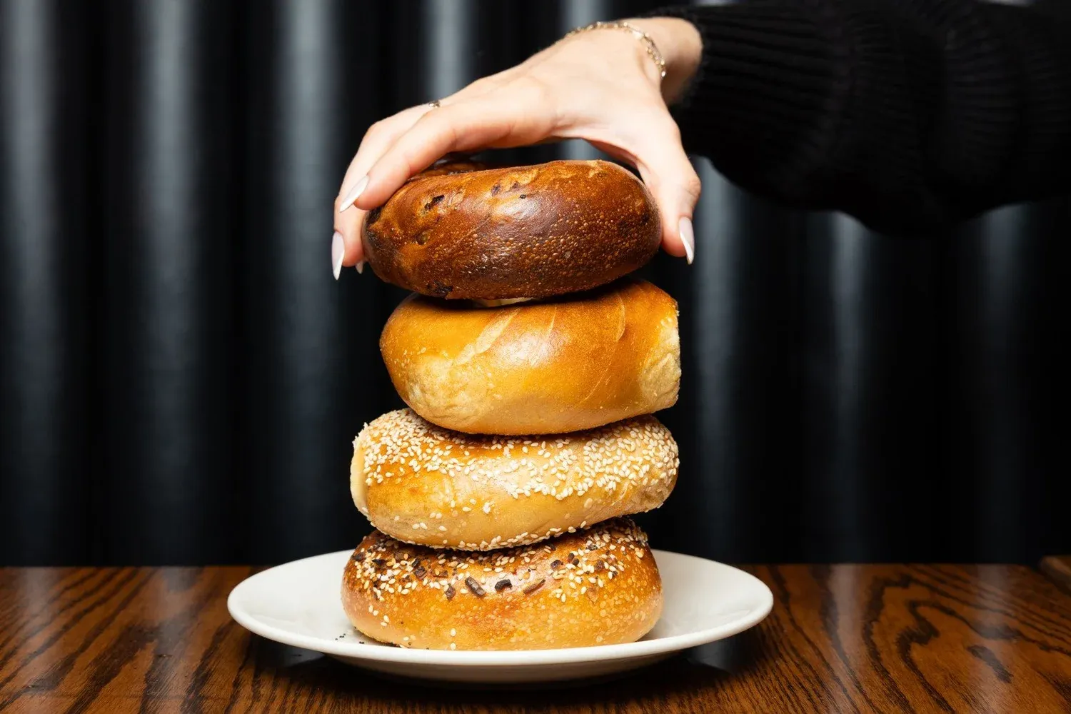 Hand pressing down on a stack of four bagels on a white plate in front of dark curtains