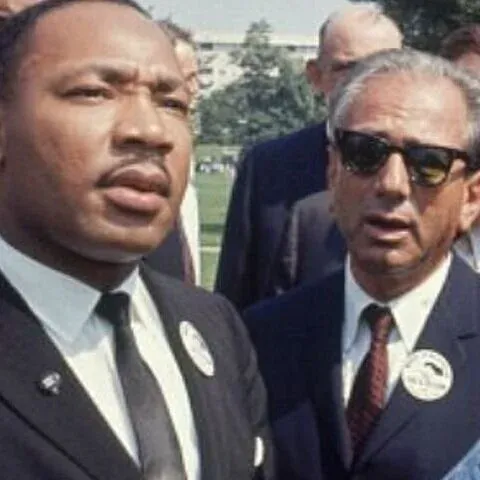 Archival color photo of Martin Luther King Jr. in a dark suit outdoors beside a man in sunglasses