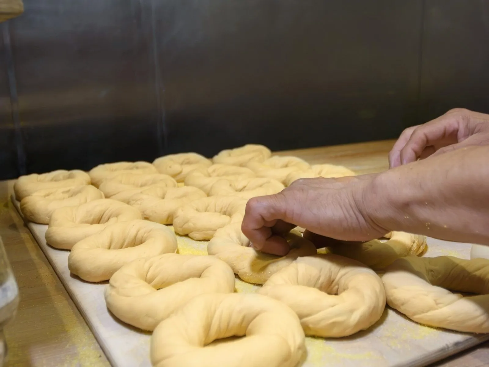 A baker's hands shaping hand-rolled bagel dough rings on a cornmeal-dusted board