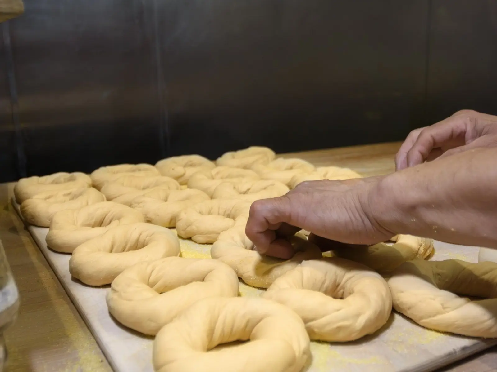 A baker's hands shaping hand-rolled bagel dough rings on a cornmeal-dusted board