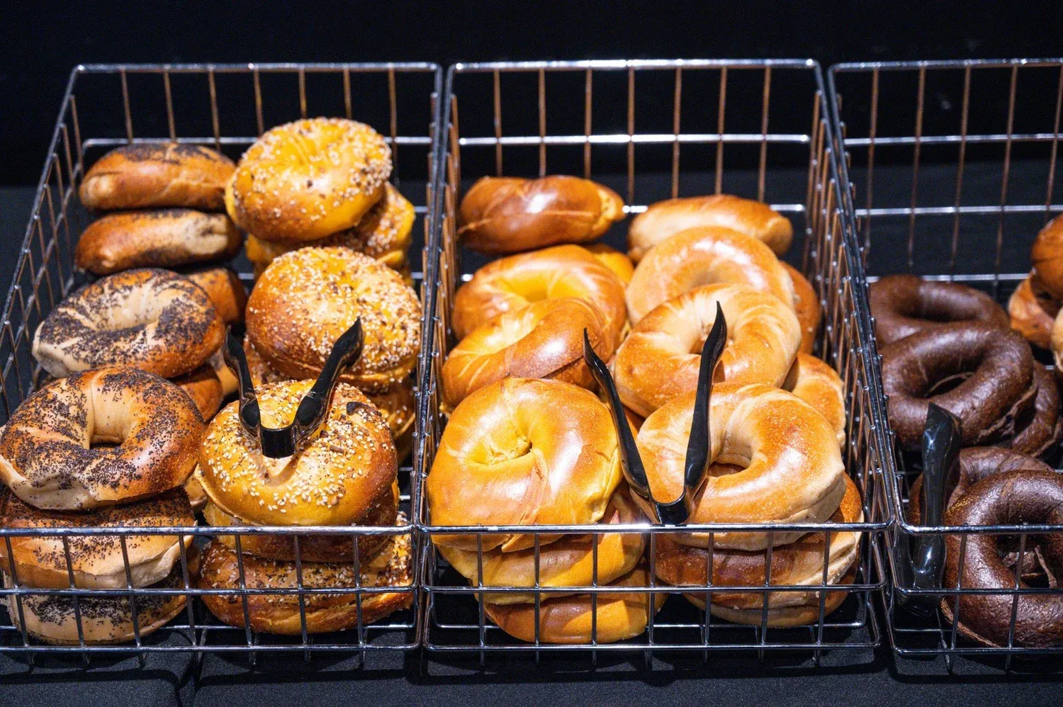 Wire baskets with serving tongs holding stacks of poppy, everything, plain and pumpernickel bagels on a black tablecloth