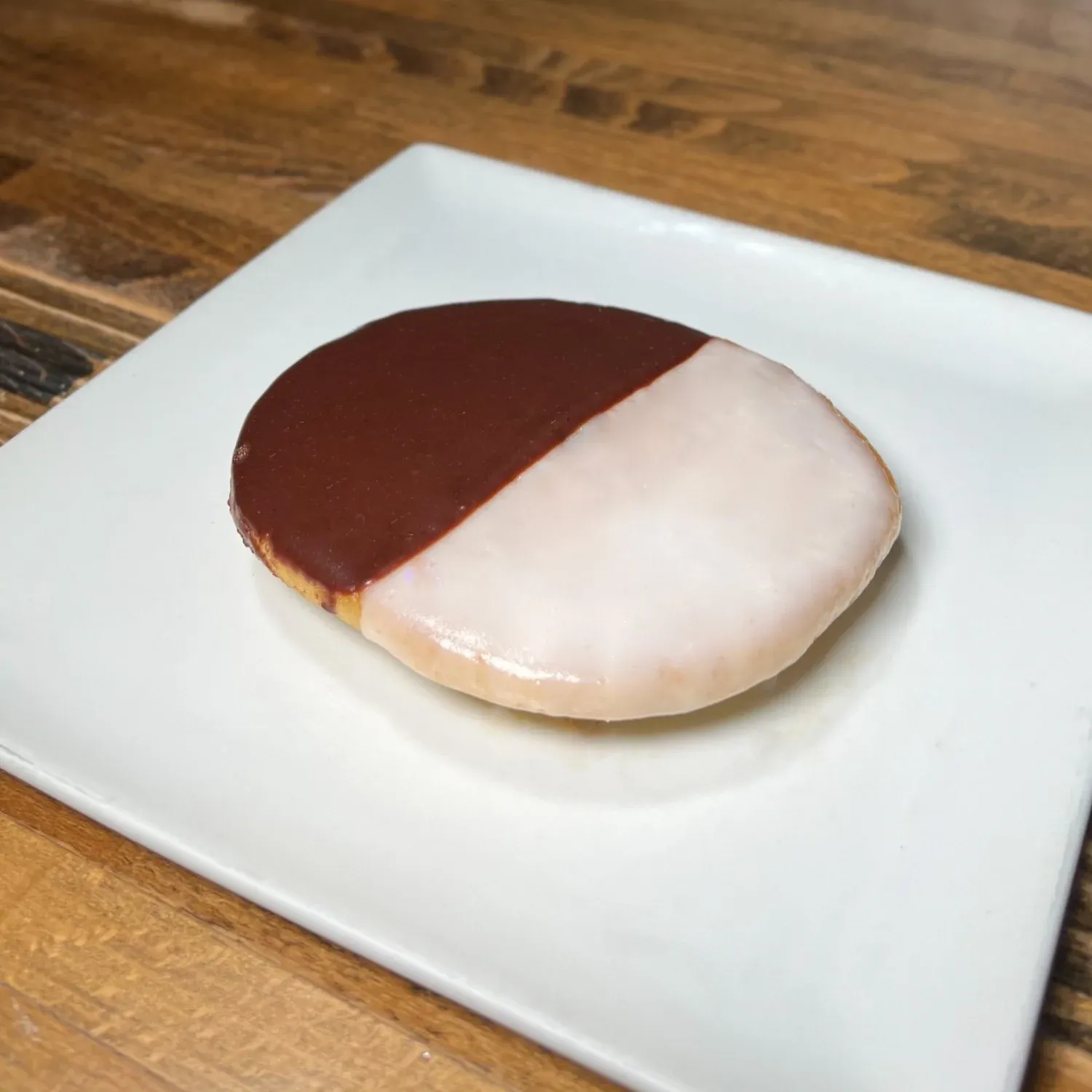 Black and white cookie, half chocolate and half vanilla icing, on a square white plate on a wooden table