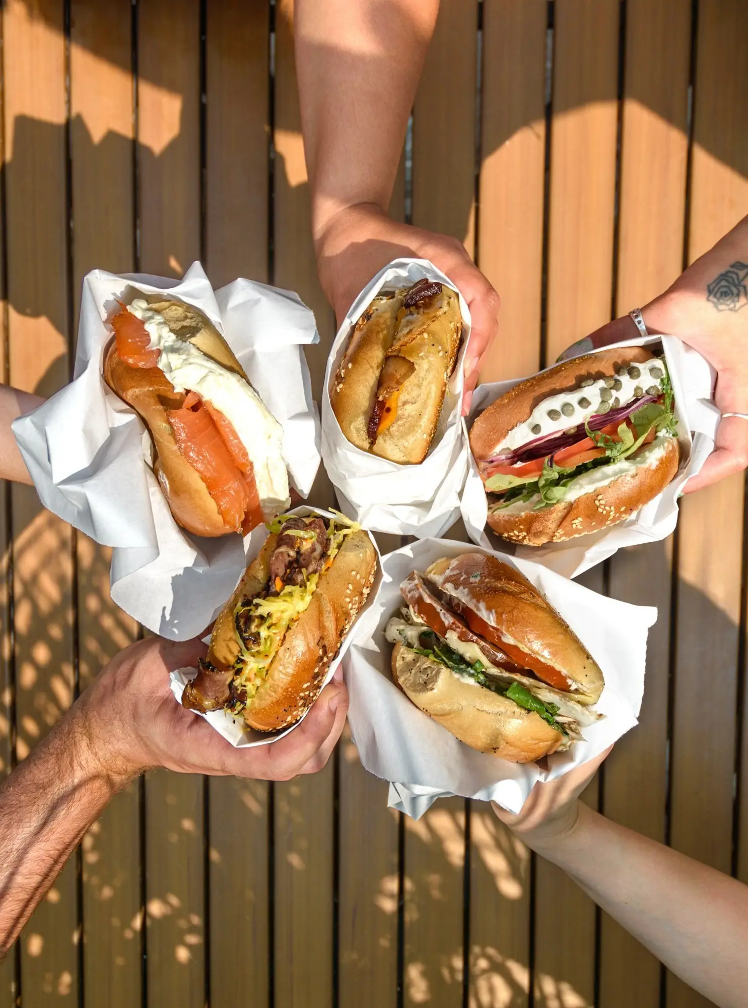 Five hands holding five wrapped Gotham bagel sandwiches in a circle, seen from above on a sunlit deck