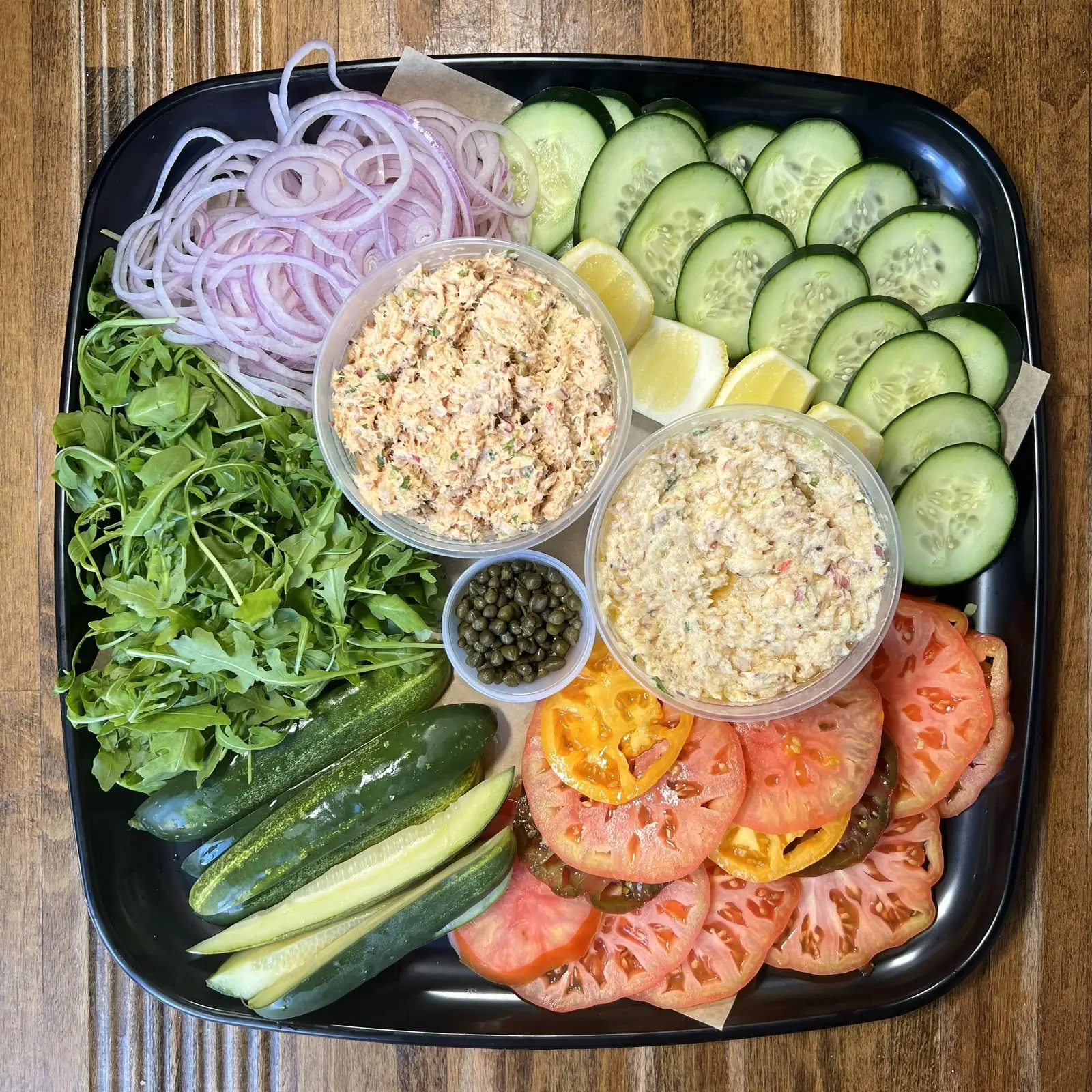 Westchester Platter from above: two deli salads with heirloom tomatoes, cucumber, arugula, pickles, red onion, capers and lemon on a black tray
