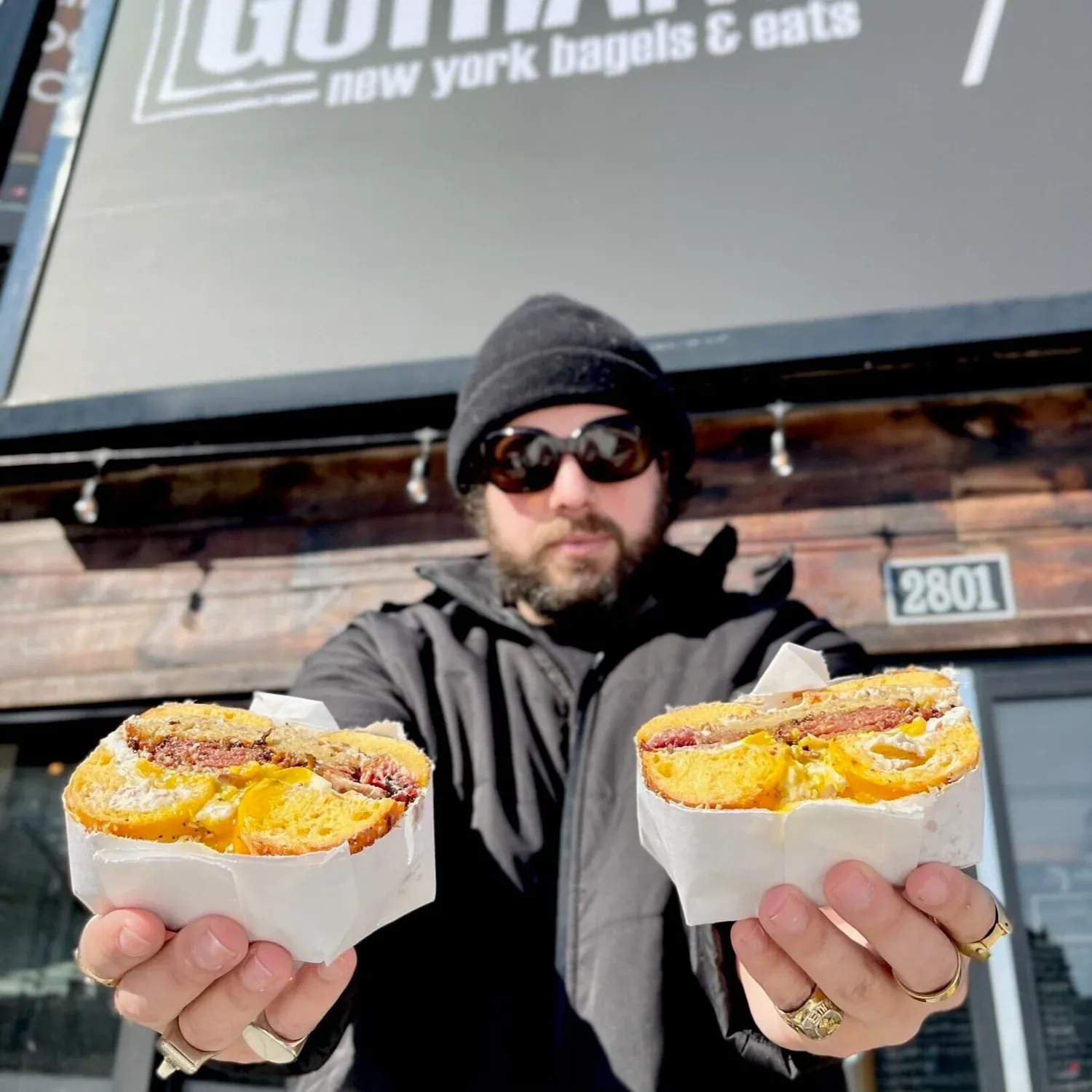 Man in a beanie and sunglasses holding two halves of an egg and meat bagel sandwich under a Gotham sign