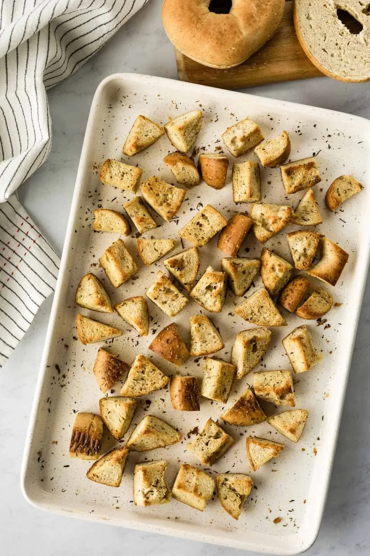 Baking sheet of toasted bagel croutons beside a striped towel and a whole and halved bagel on a cutting board