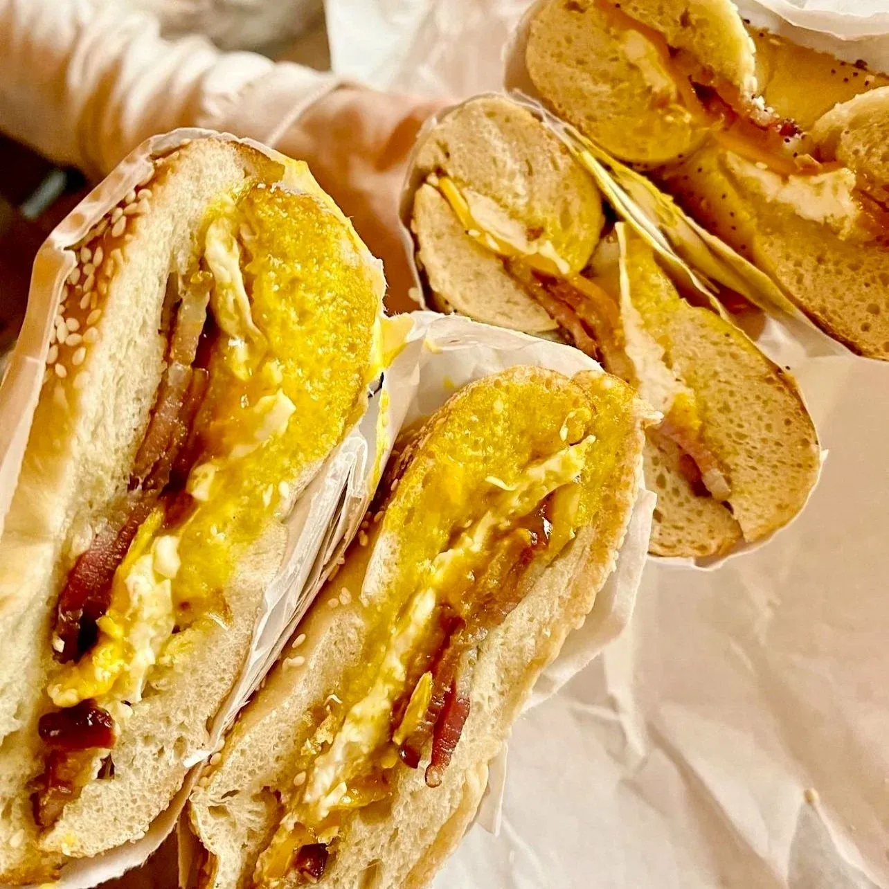 Halved sesame bagel sandwiches with fried egg, bacon and melted cheese wrapped in white paper