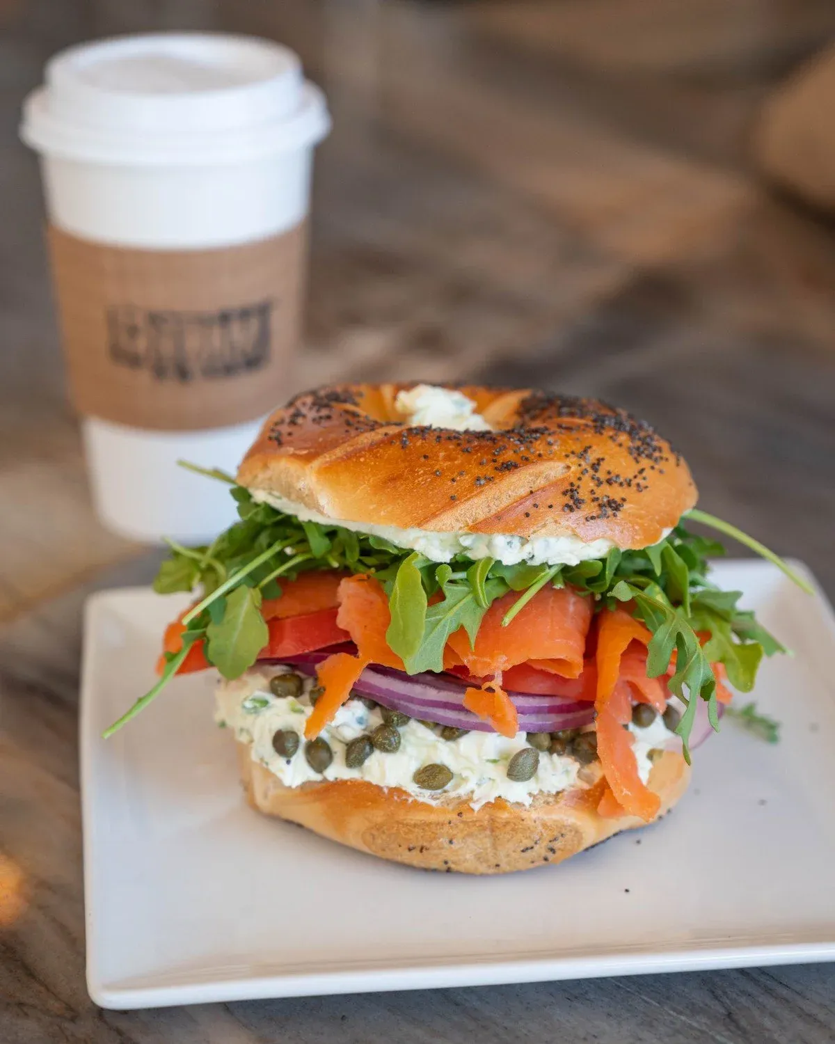 Poppy seed bagel stacked with smoked salmon, red onion, arugula, capers and herbed cream cheese beside a paper coffee cup