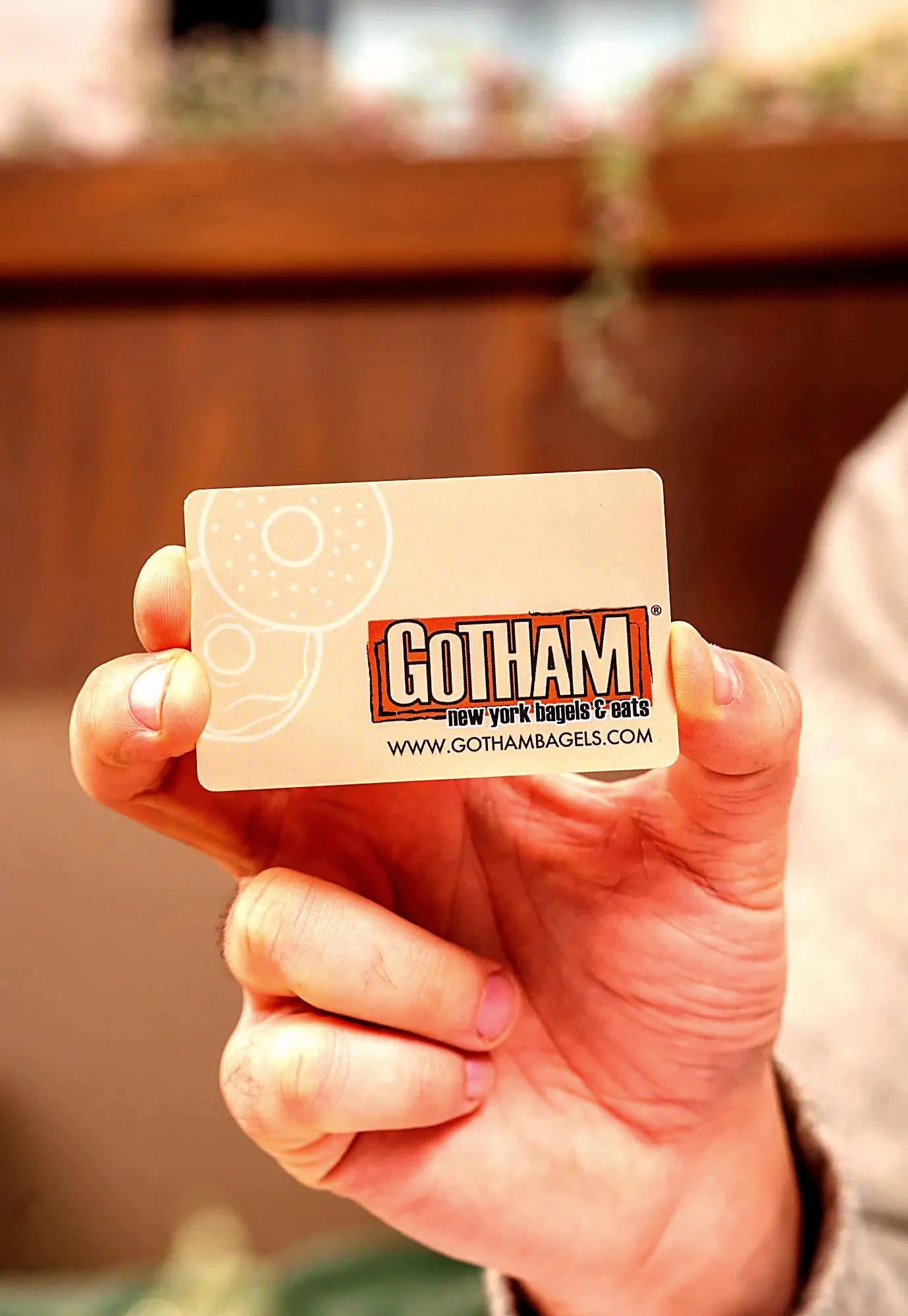 A hand holding up a cream Gotham Bagels gift card with the orange Gotham logo