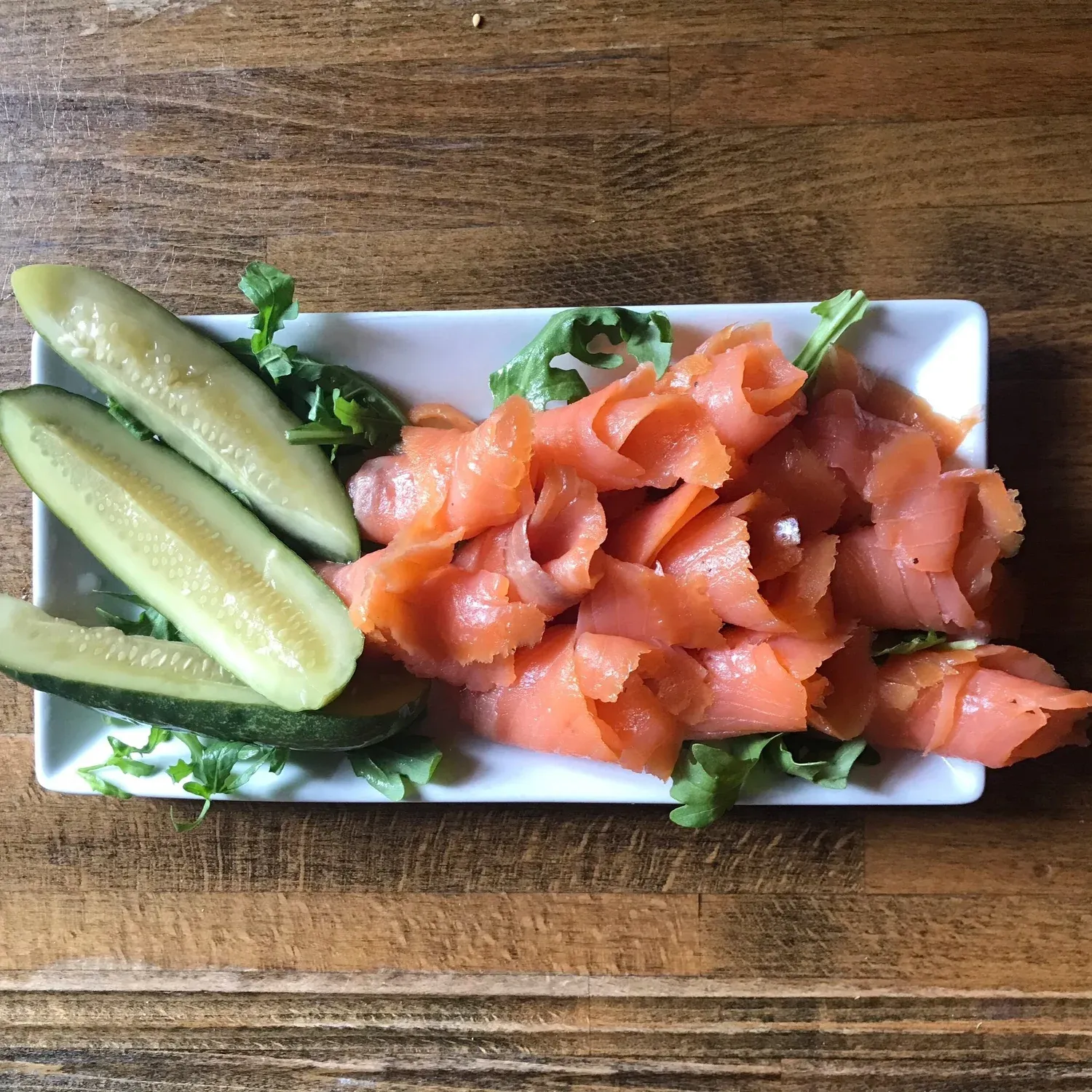 Folded slices of smoked salmon with pickle spears and arugula on a rectangular white plate