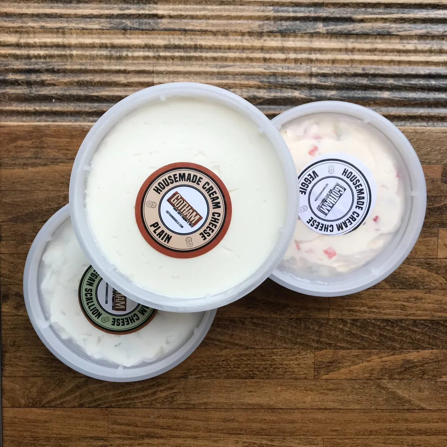 Three tubs of Gotham housemade cream cheese labeled plain, veggie and vegan scallion on a wooden counter
