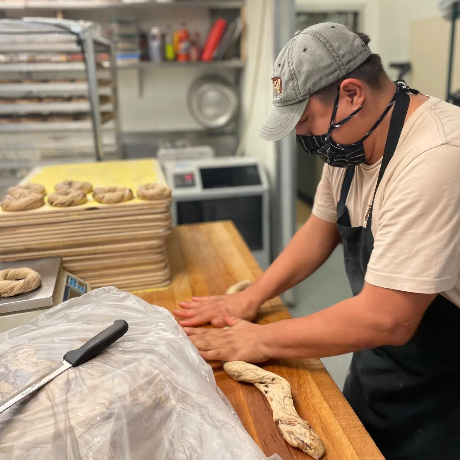 Masked baker in a cap and apron rolling bagel dough by hand on a wooden bench, shaped bagels behind