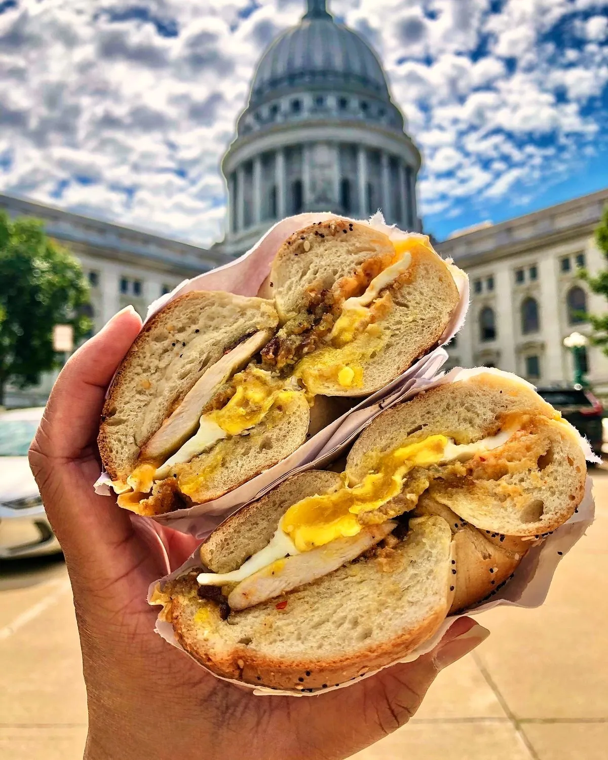A Gotham Madison egg sandwich on a hand-rolled bagel, held up in front of the Wisconsin State Capitol