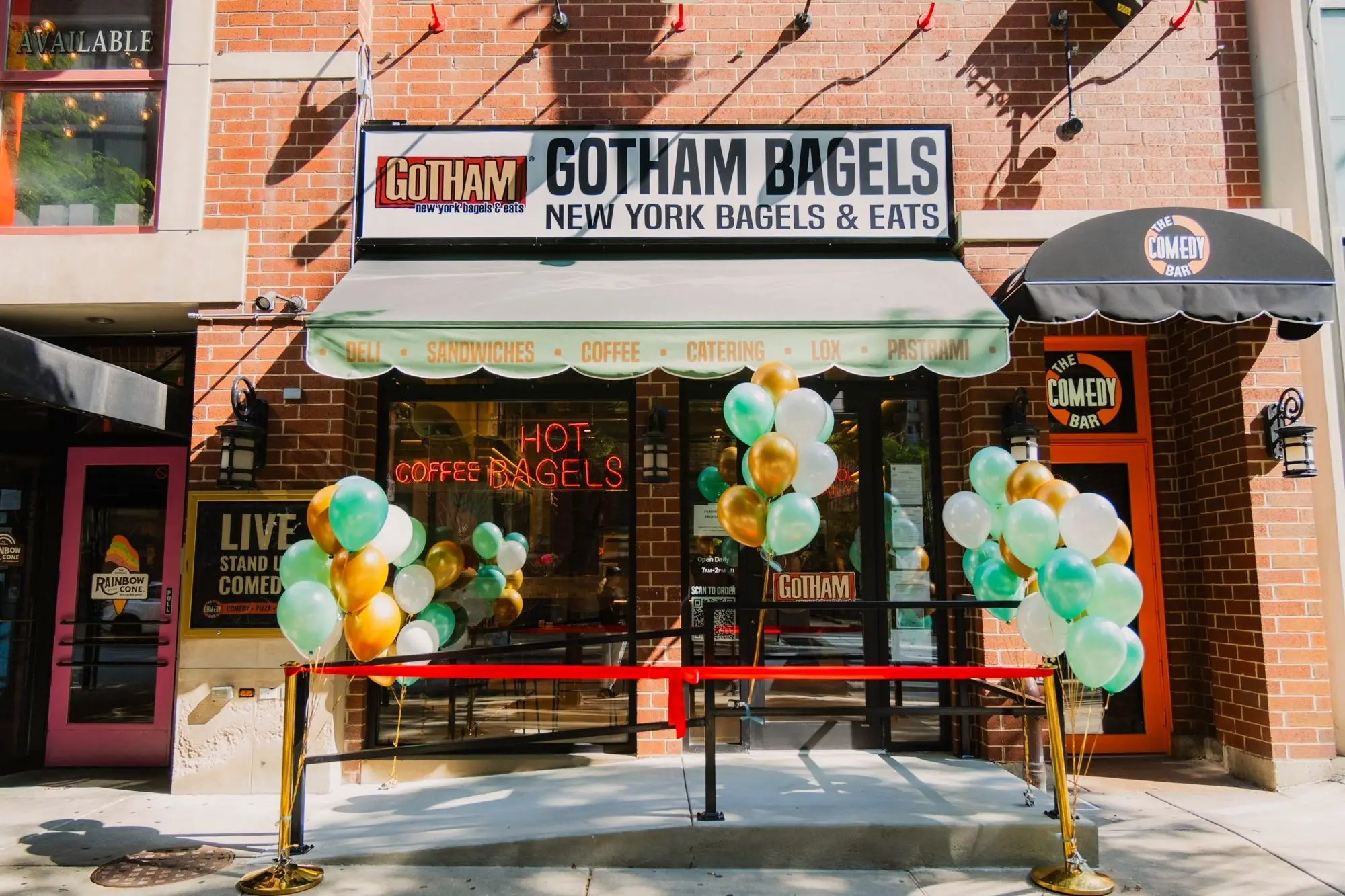 Gotham Bagels Magnificent Mile storefront on opening day, with green and gold balloons and a red ribbon across the entrance under the deli, sandwiches, coffee, catering awning