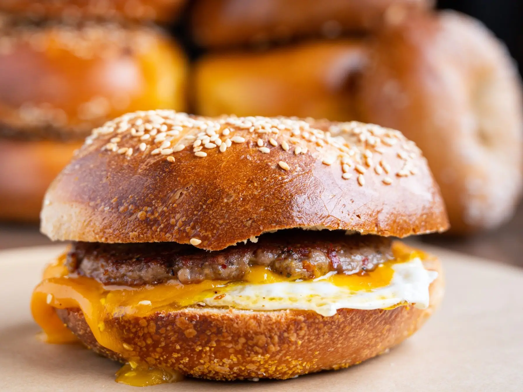 Spicy sausage, egg and cheddar on a sesame bagel, the cheddar melting over the egg, with stacks of bagels behind