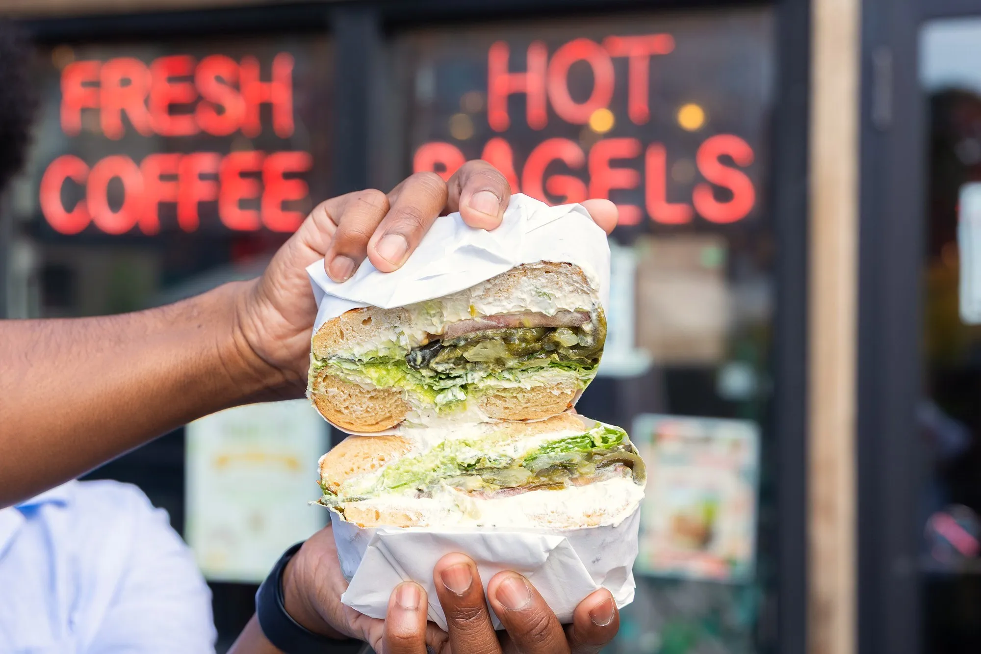 A wrapped bagel sandwich, cut in half and held up in front of Fresh Coffee and Hot Bagels neon