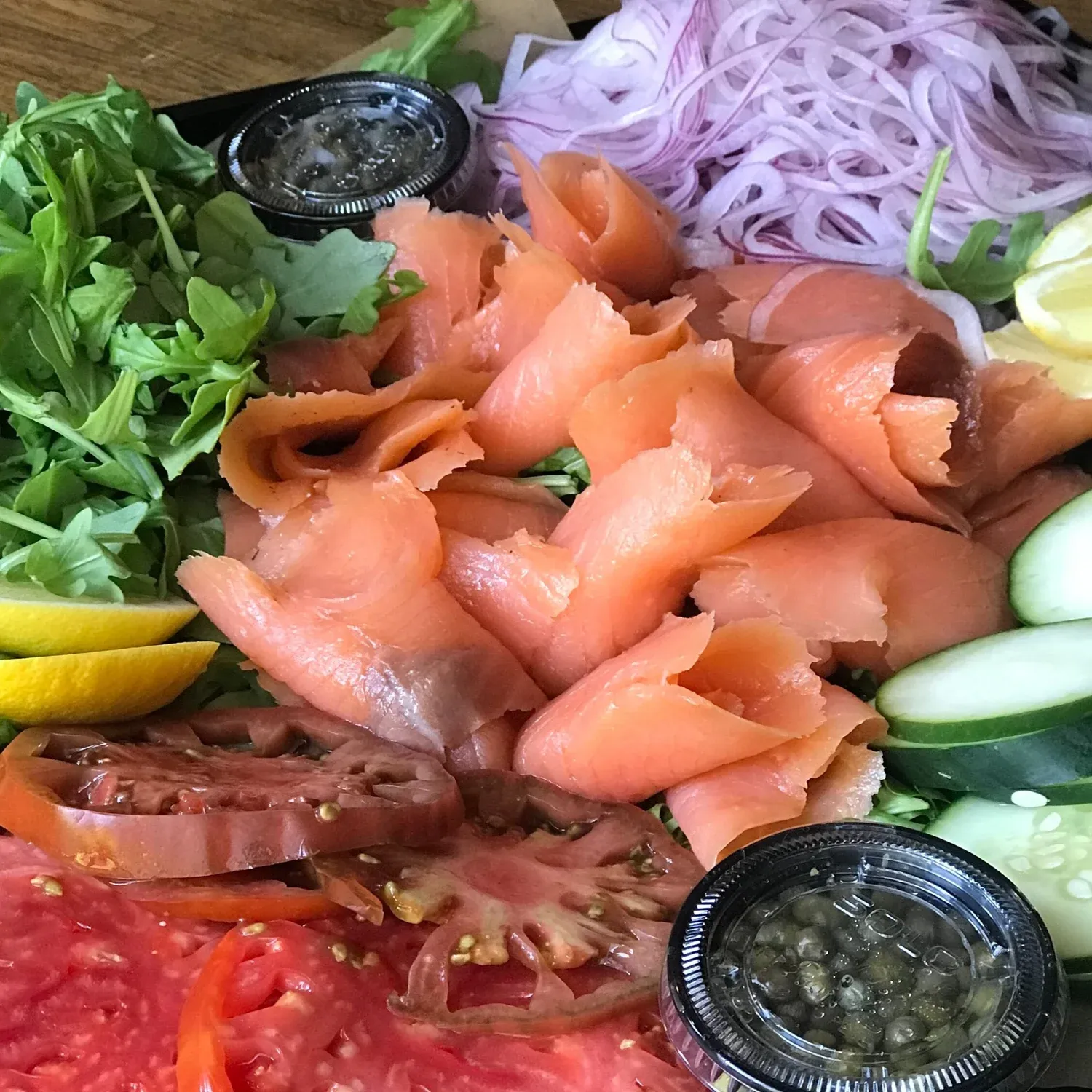 Platter of smoked salmon with arugula, shaved red onion, lemon wedges, tomato slices, cucumber and capers