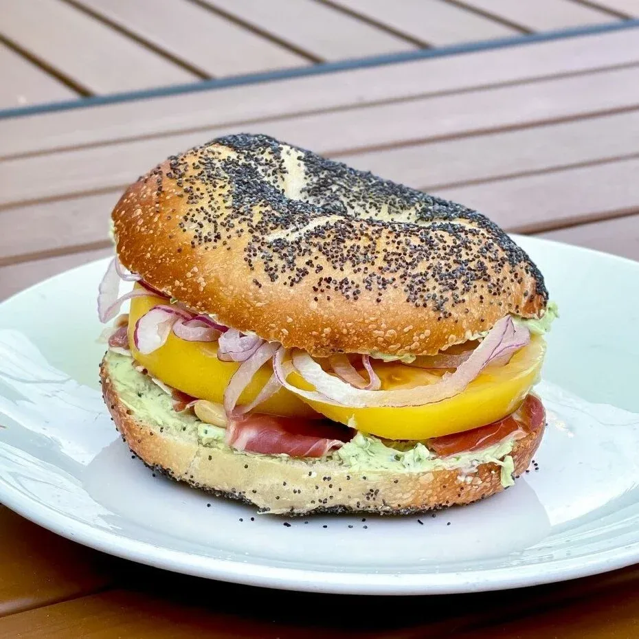 Poppy seed bagel sandwich with yellow tomato, red onion, prosciutto and herbed cream cheese on a white plate outdoors