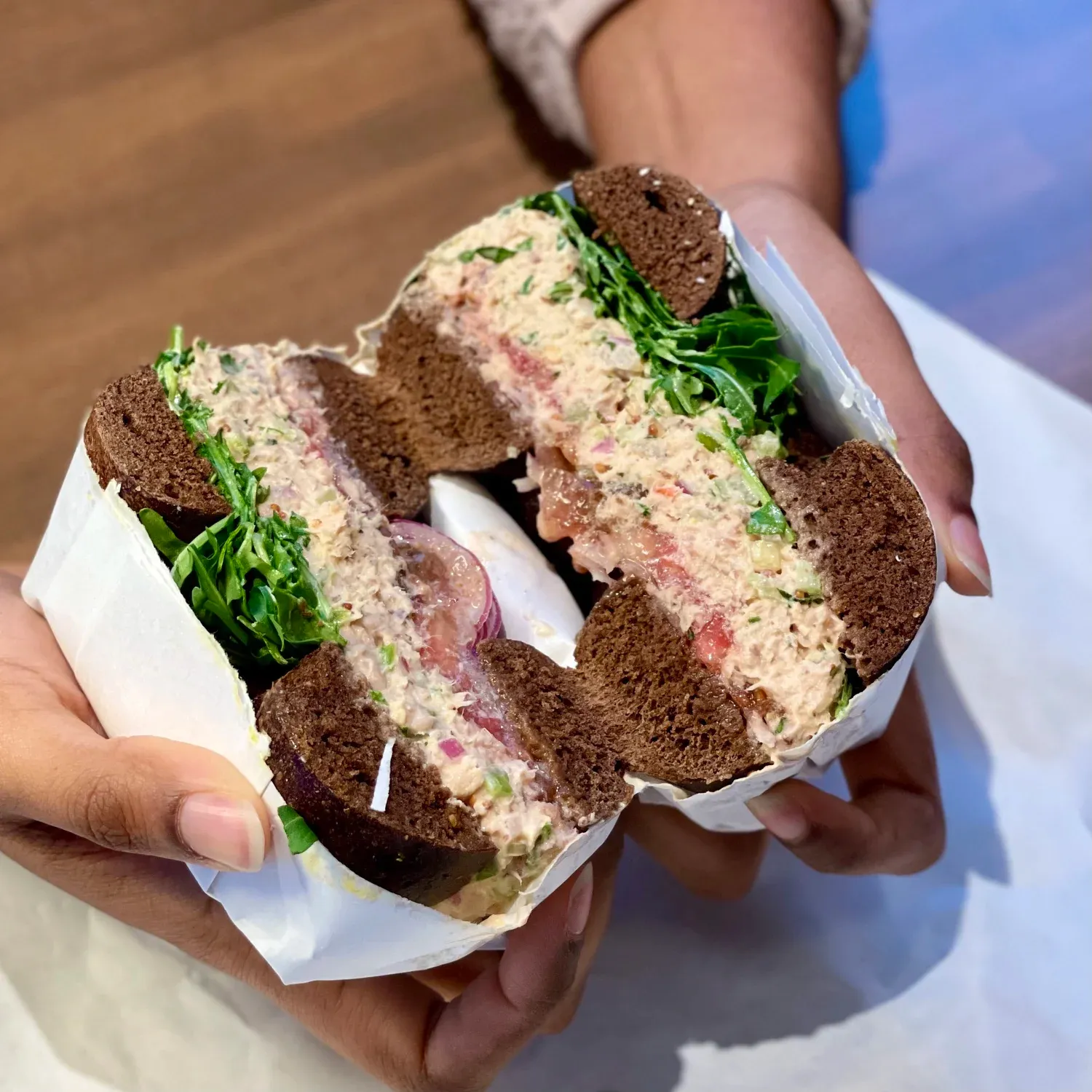 Hands holding a pumpernickel bagel sandwich cut to show tuna salad, tomato, red onion and arugula in paper