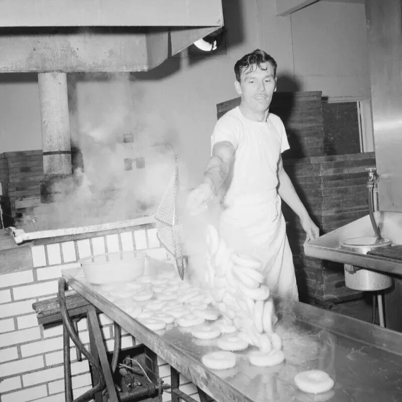 Black-and-white photo of a baker lifting boiled bagels from a steaming kettle with a wire basket