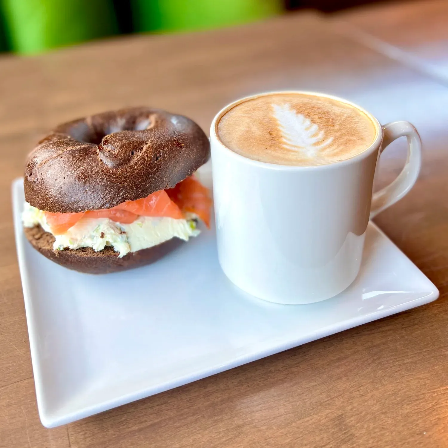 Latte with fern-shaped foam art in a white mug beside a pumpernickel bagel with scallion cream cheese and smoked salmon