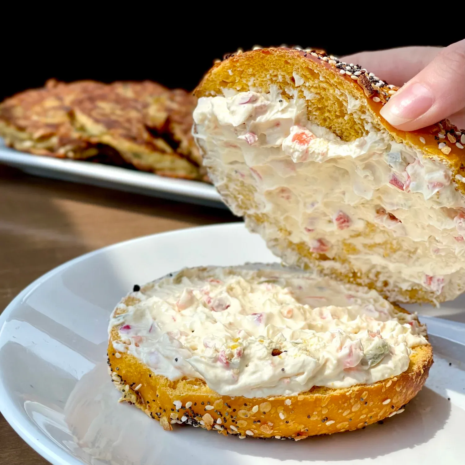 Hand lifting the top of an everything bagel spread thick with veggie cream cheese, bottom half on a white plate