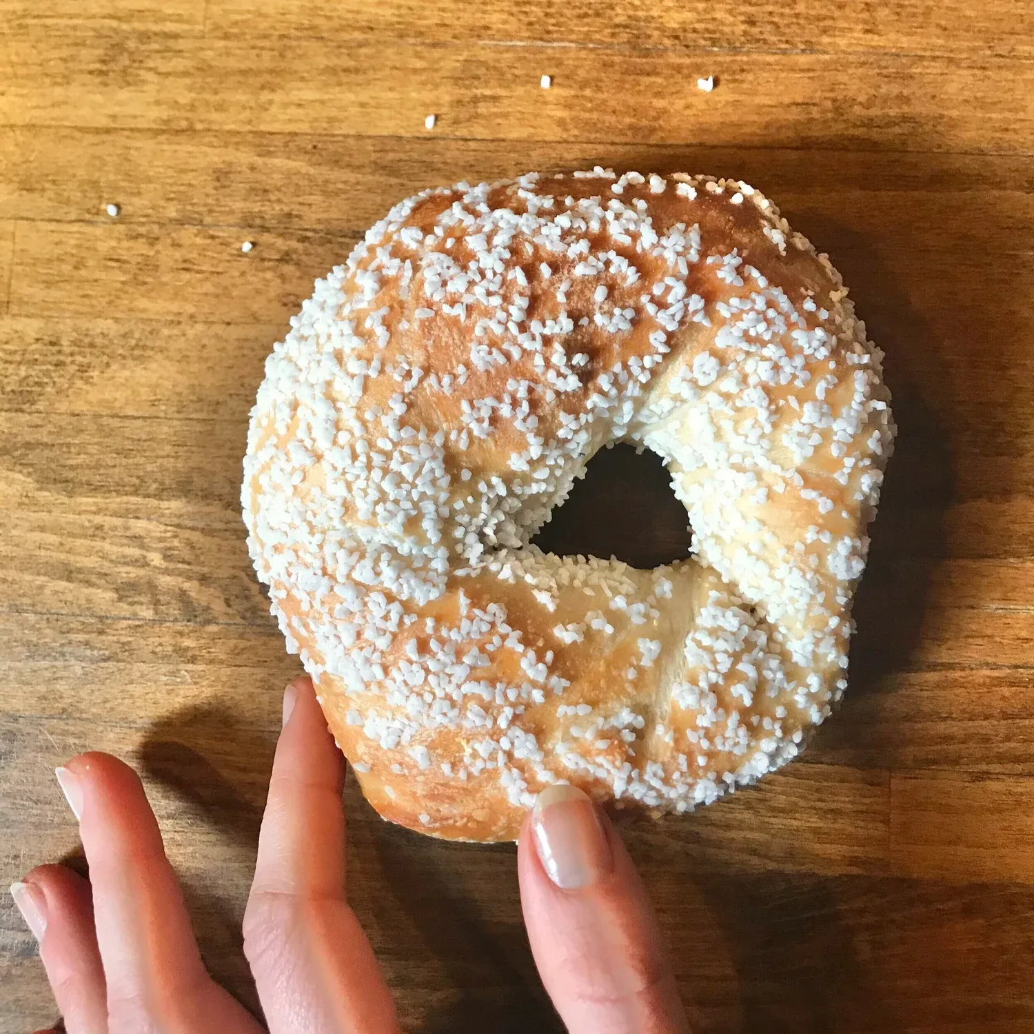 Fingers holding a bagel crusted with coarse white salt crystals on a wooden table