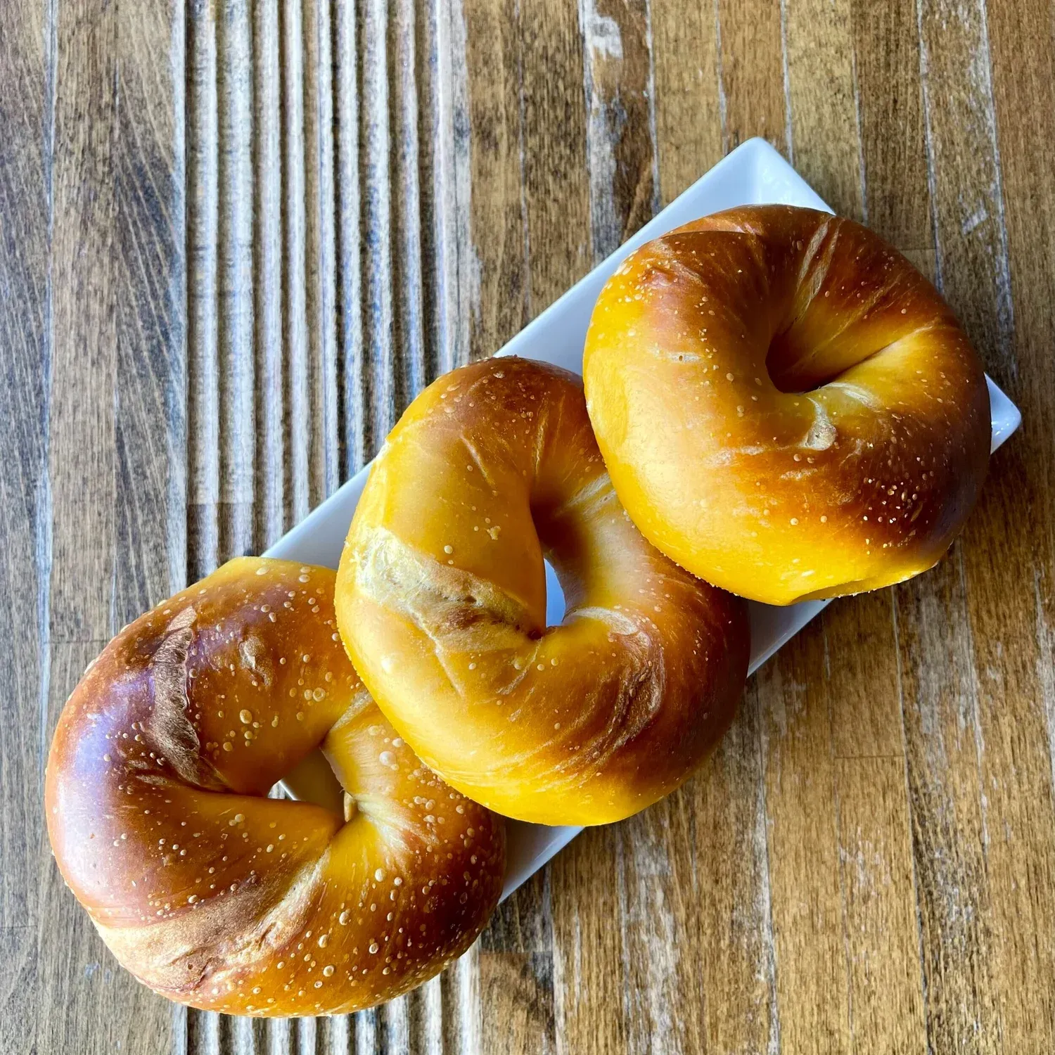 Three glossy golden egg bagels on a white rectangular plate on a wooden table