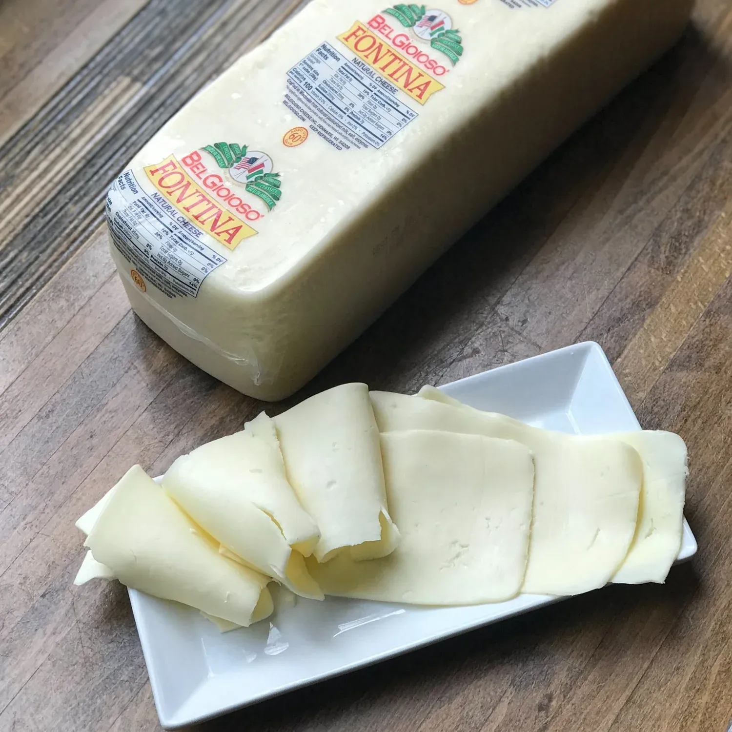 Folded slices of fontina on a white plate in front of a large labeled block of BelGioioso fontina