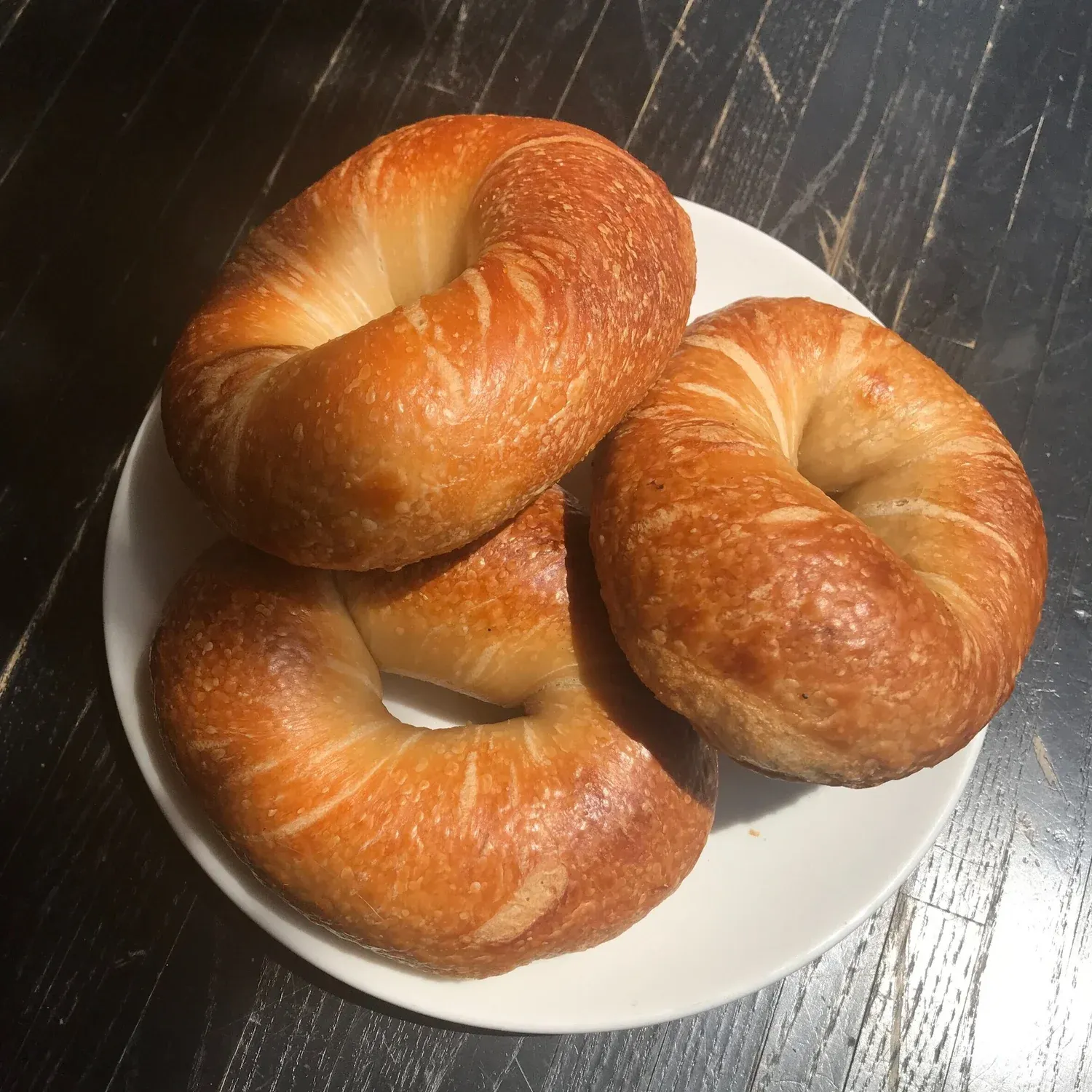 Three glossy plain bagels stacked on a white plate on a dark wooden table in sunlight