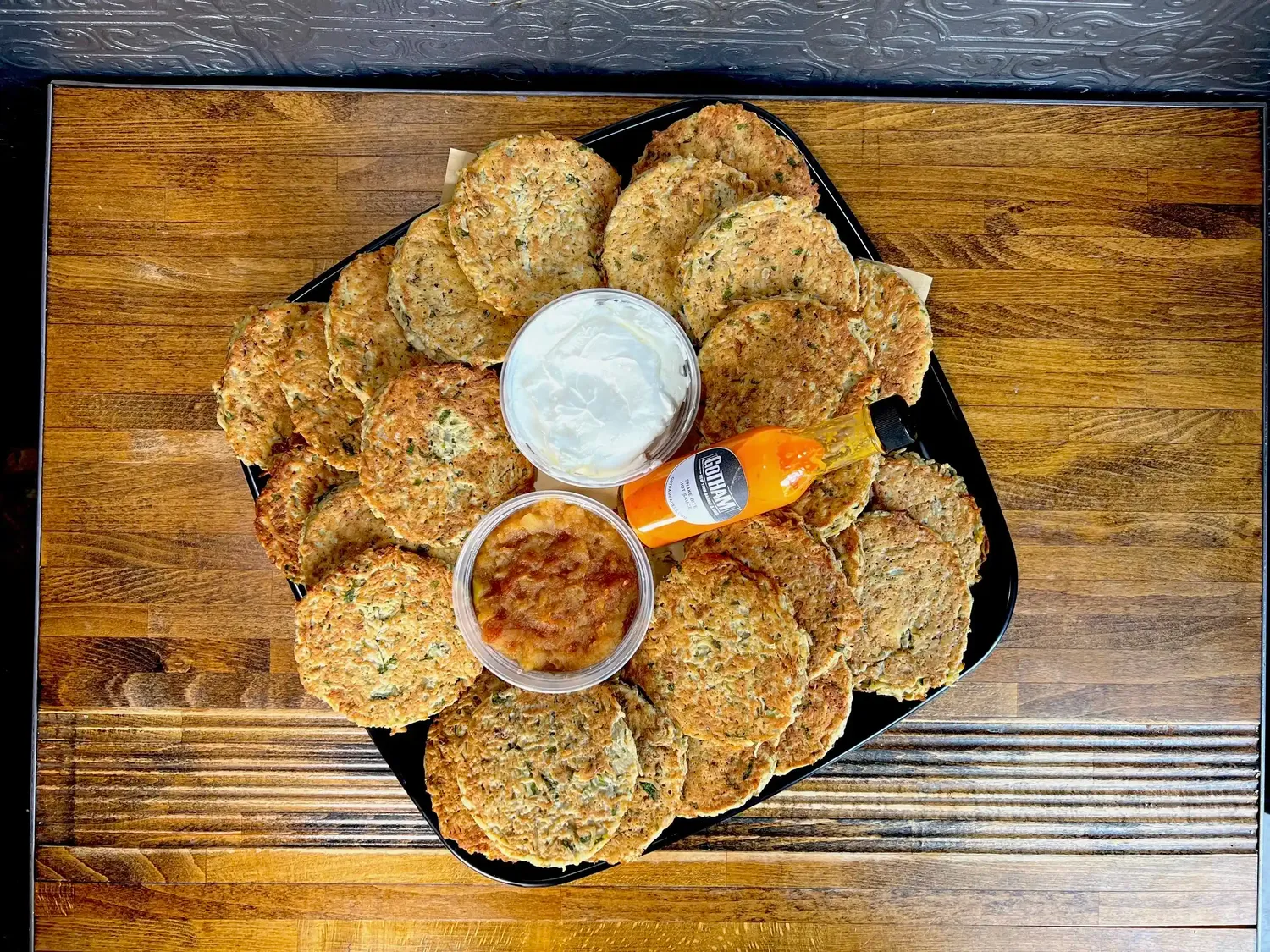 Catering tray of golden potato latkes around tubs of sour cream and applesauce and a bottle of Gotham hot sauce