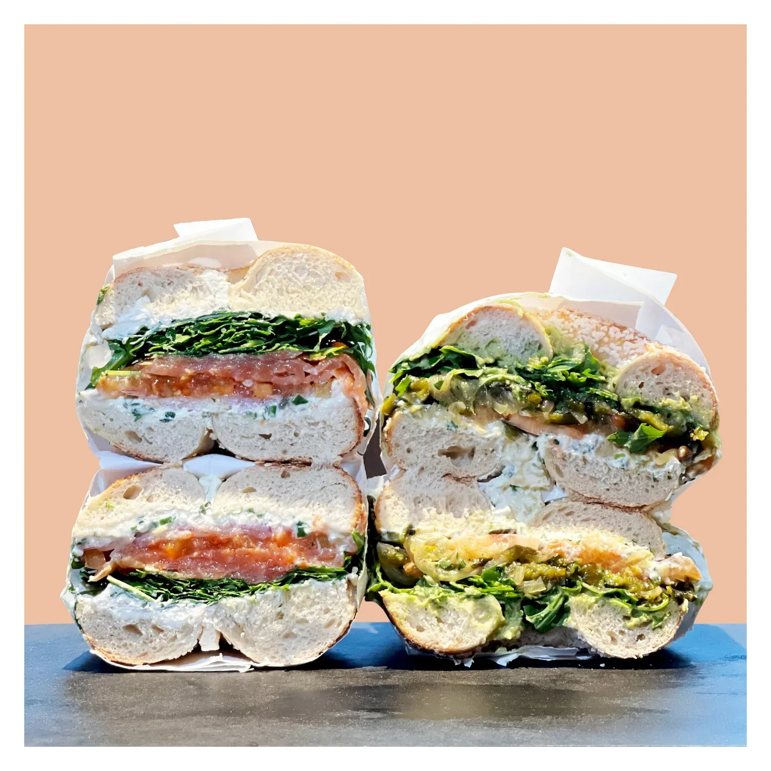 Two halved bagel sandwiches stacked in paper, one with smoked salmon and greens, one with guacamole and arugula